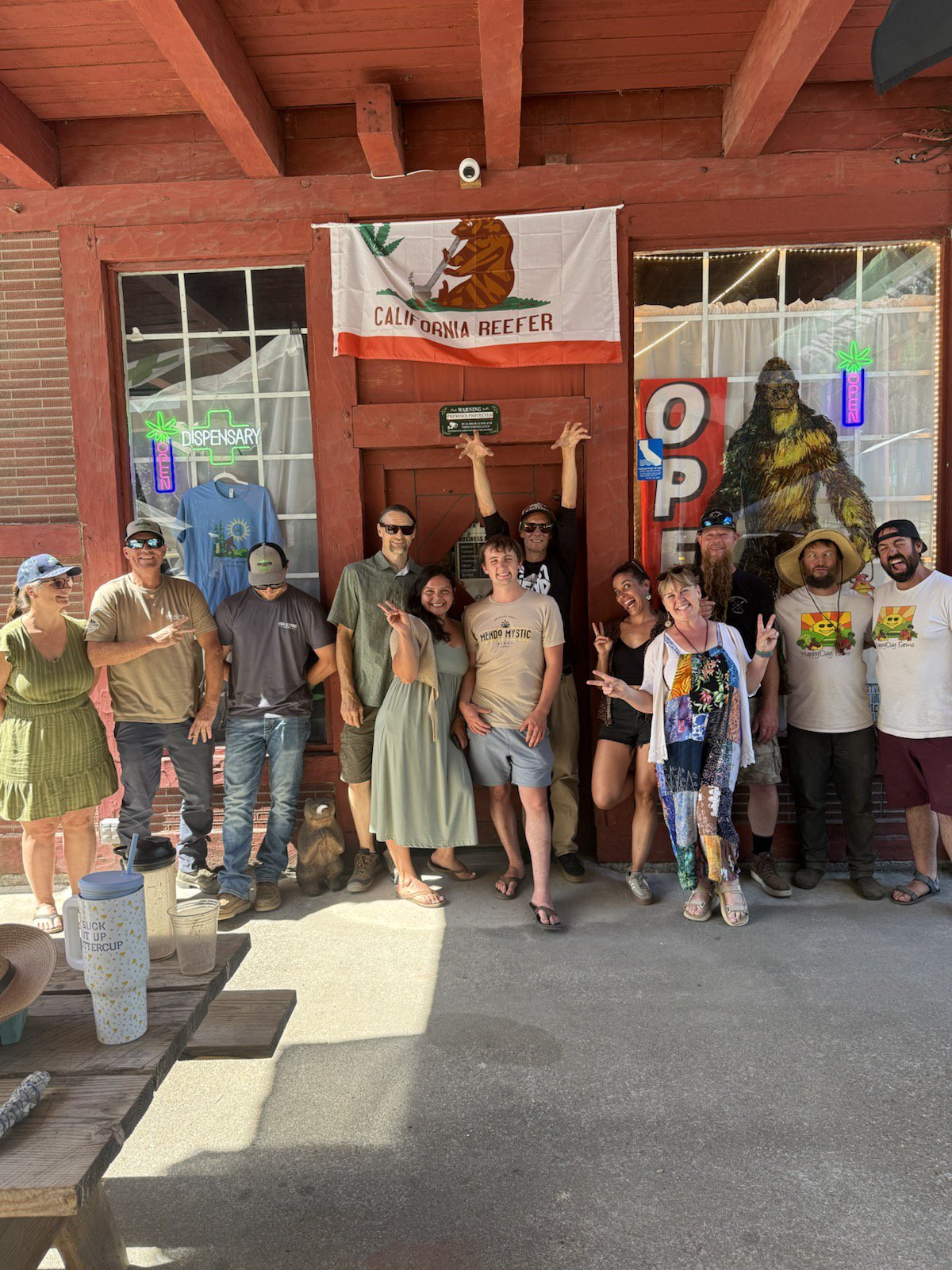 A group of twelve people stand together in front of the red wooden storefront of Emerald Sasquatch at the Peg House in Leggett, California. Above them hangs a flag that says California Reefer featuring a bear. The group is smiling and making peace signs and playful hand gestures. Some individuals wear shirts representing local farms like Mendo Mystic and Happy Day Farms. To the right, a large illustration of a Sasquatch is visible in the window next to a neon open sign. The scene is outdoors on a sunny day with a wooden picnic table in the foreground.